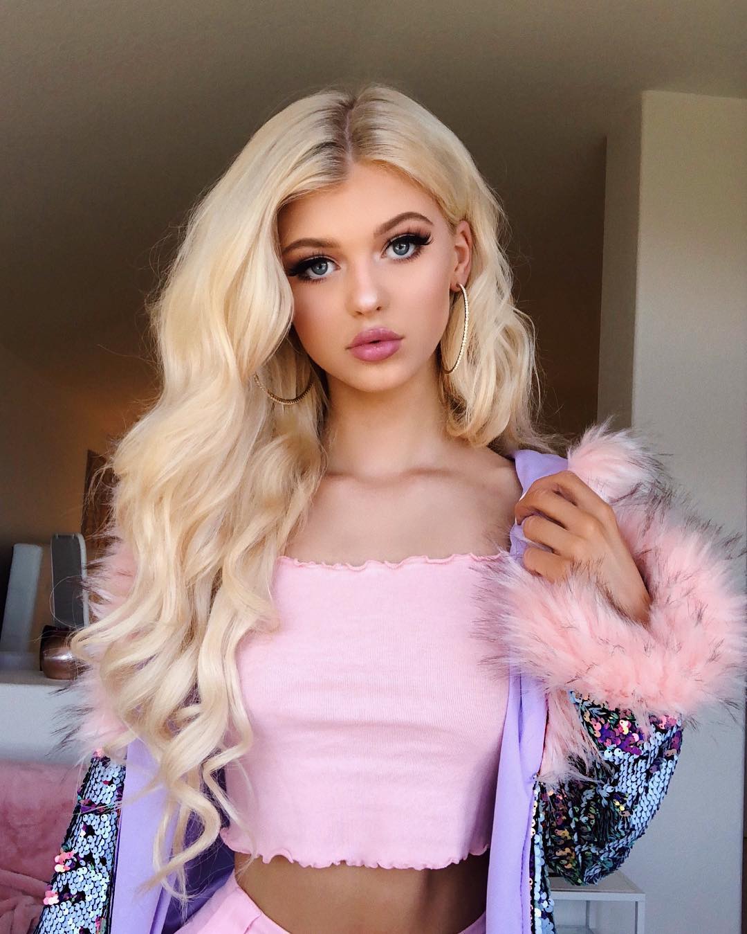 a blonde woman with blue eyes is posing indoors. she has heavy makeup and is wearing large hoop earrings. she's dressed in a pink crop top and a sequined jacket with pink fur trim. she is holding the jacket open with one hand.