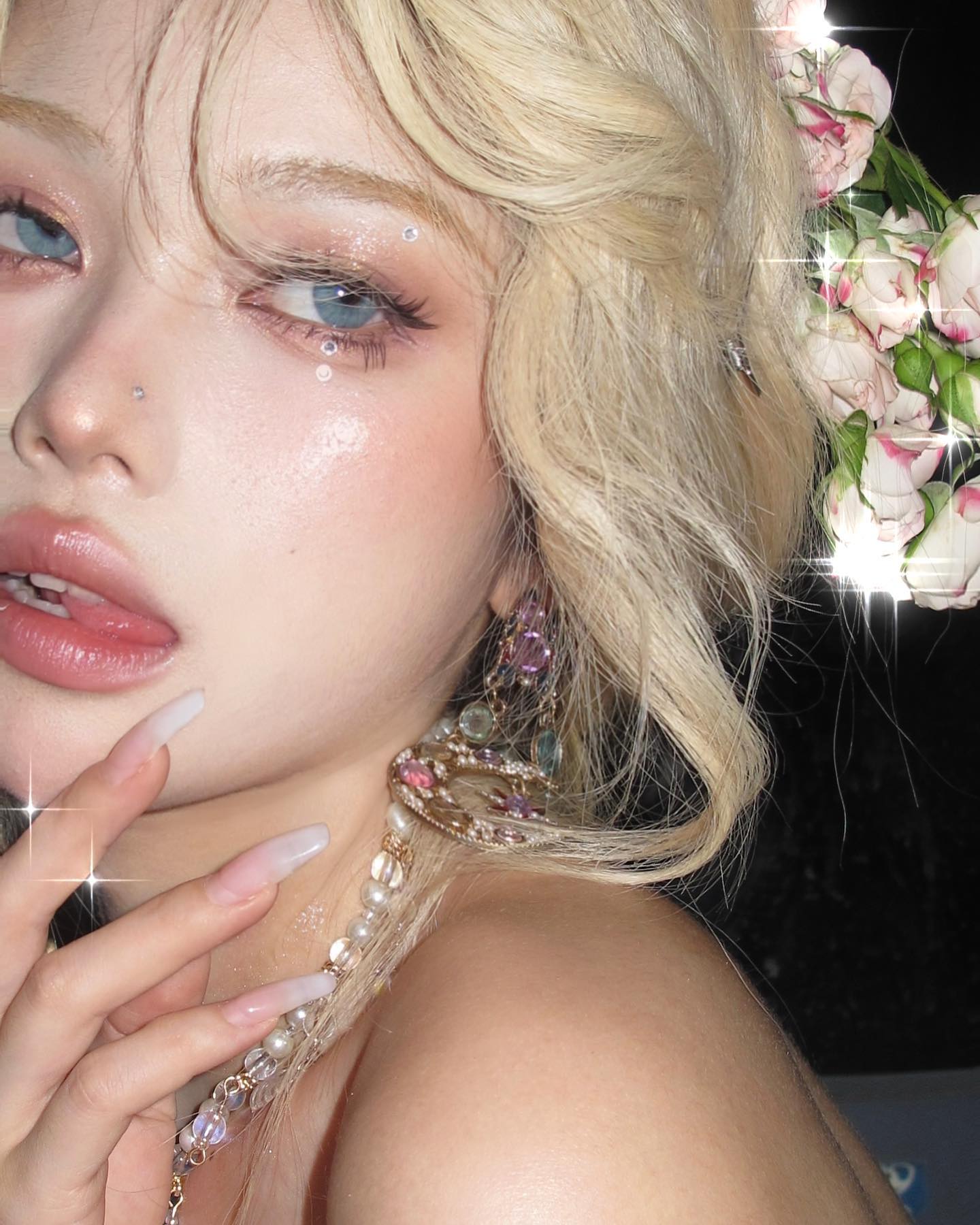a closeup selfie portrait of a young woman with blonde hair. she has on makeup that emphasizes a soft, ethereal aesthetic with rosy hues and glitter. she is wearing elaborate earrings and a necklace with pearls and colorful gems. she has a flower crown resting on her head.  the overall impression is feminine and romantic.