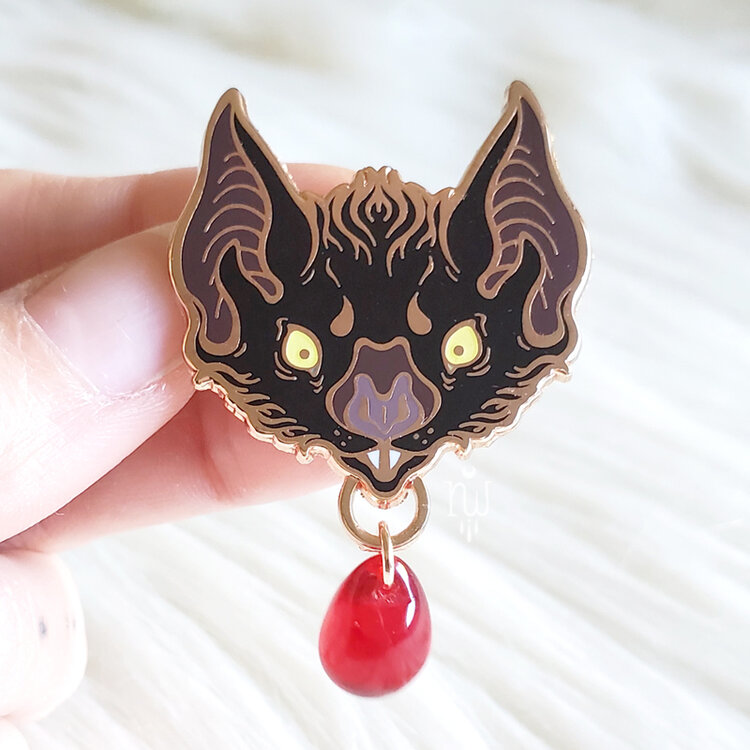 a hand holds a batshaped enamel pin with a red gem charm dangling from the bottom. the pin is black with bronze edges and a purple nose. it has large, pointed ears and a red gem. the background is a soft white material.