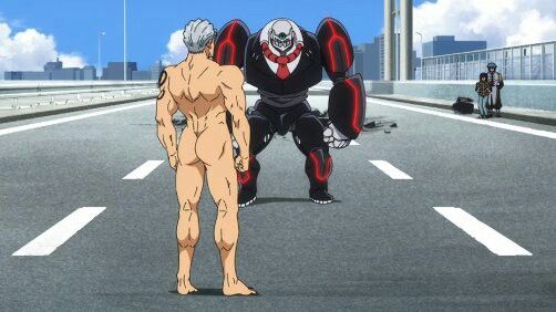 a nude man with gray hair and a tribal tattoo on his back is standing in the middle of a city street, facing a large robot. the robot is black and red with a white skulllike face. in the background, there are two figures standing on the sidewalk, seemingly unfazed by the confrontation. the scene is set during the day with clear skies.