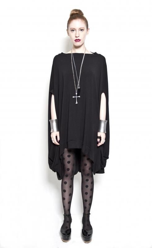 a woman stands against a white backdrop, facing forward. she is dressed in a long, black ponchostyle dress that reaches above the knees. the dress has slits in the sleeves near the shoulders. she is also wearing black tights with polka dots, and shiny black platform heels. the woman is wearing a silver cross necklace, and matching silver cuffs on her wrists. her hair is styled in a bun on top of her head.