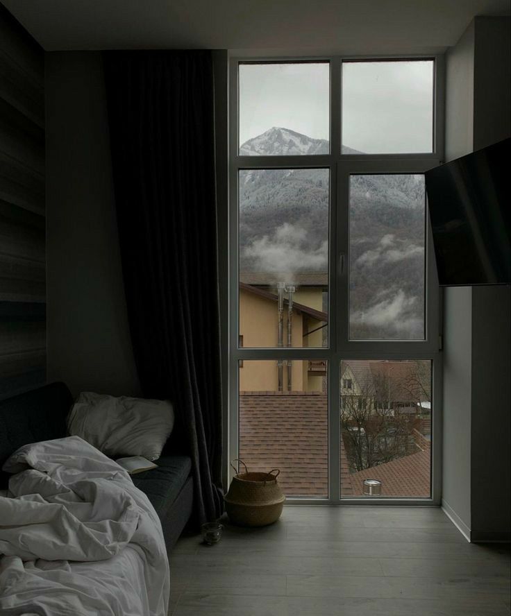he image shows a cozy room with a large window overlooking a snowy mountain. the room is decorated in a dark and moody style, with grey walls, dark curtains, and a simple bed and couch. a basket sits on the floor near the window, and the overall feeling is one of calm and serenity.