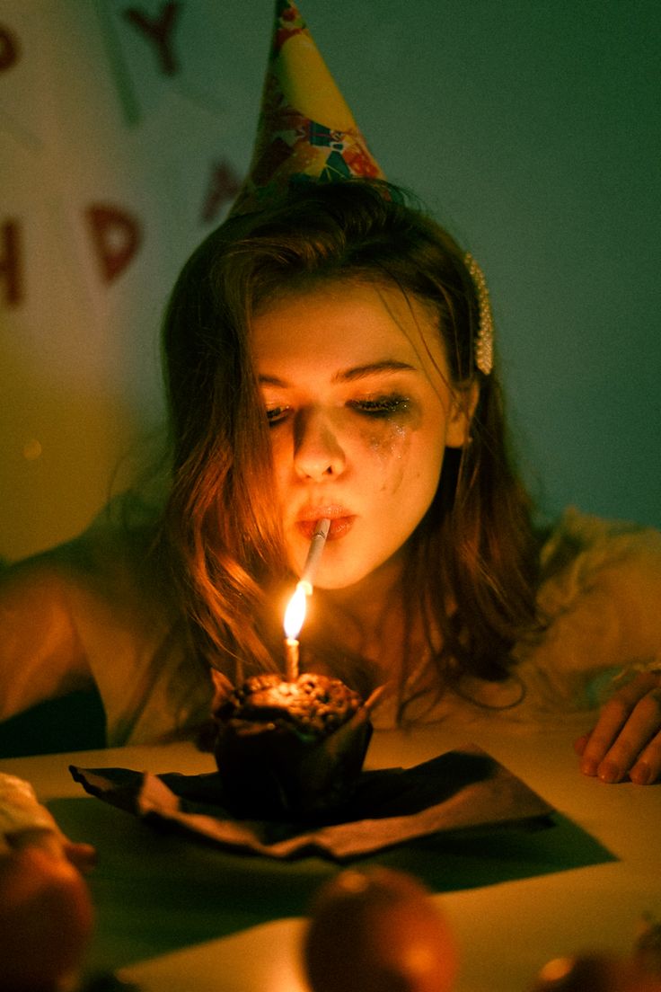 a young woman wearing a party hat sits alone at a table, looking sad and forlorn.  she's crying and smoking a cigarette, while a lit candle sits atop a cupcake in front of her. the lighting is dim and moody, enhancing the somber atmosphere. the overall image conveys a sense of loneliness and emotional turmoil on a birthday.