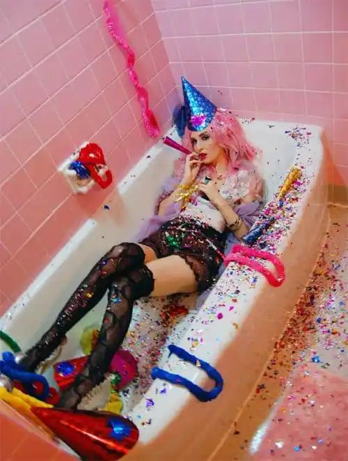 a young woman in a sparkly dress and a party hat is sitting in a bathtub surrounded by confetti. she is smiling and looks like she's having a lot of fun. the image is full of color and joy, and it's a great example of how to celebrate a special occasion.