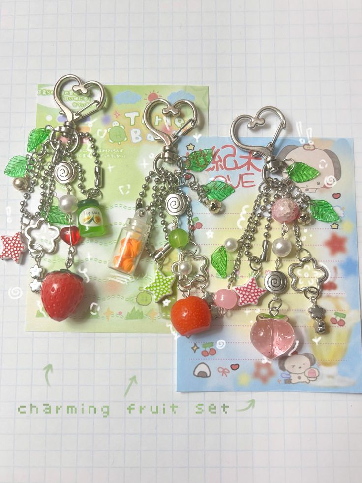 three keychains with a variety of fruit and decorative charms are displayed against pastelcolored backgrounds. each keychain has a heartshaped clasp and features a different fruit charm: a strawberry, a tangerine, and a peach. the keychains are adorned with beads, miniature bottles, and starshaped, leafshaped, and flowershaped charms. the objects are set on top of cards with illustrations. the background is a graph paper. the caption "charming fruit set" is at the bottom of the frame.