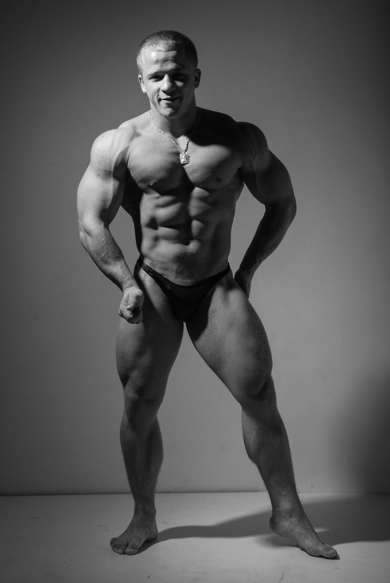 a black and white fulllength studio shot of a muscular male bodybuilder. he is posing, showcasing his welldefined physique. his muscles are highly developed, and his expression is confident. he is wearing darkcolored briefs and a necklace. the background is a simple, even gray, with subtle lighting creating shadows that emphasize his musculature. the overall image projects strength, fitness, and health.