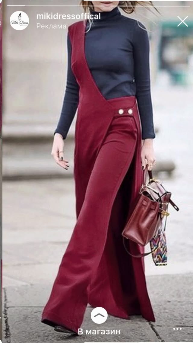a woman is standing outdoors wearing a dark blue turtleneck and a burgundy pair of pants with a draped asymmetrical overlay. the pants are widelegged and have two prominent buttons on the hip. she is carrying a burgundy handbag in her right hand. the image appears to be part of an instagram advertisement.
