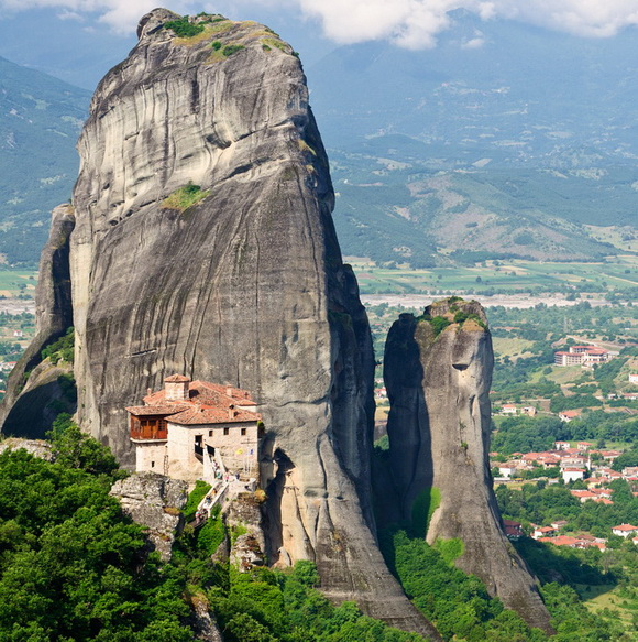 the image shows the meteora monasteries in greece. the monasteries are built on top of tall, rocky cliffs. in the foreground, a monastery is perched on a cliff with a winding stairway. the background shows a mountain landscape with a small village.