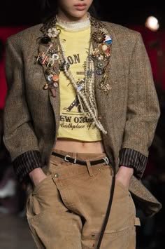 a female fashion model wearing a brown tweed blazer with various pins and patches, a yellow tshirt with a graphic print, and brown pants with a black belt. she is posing on a runway with her hands in her pockets. her style is trendy and chic.  she is wearing a lot of jewelry around her neck.