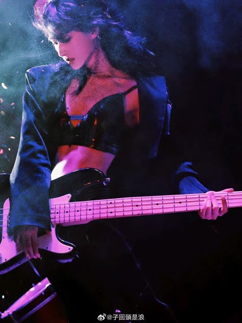 a woman is playing a bass guitar on a stage during a performance. she is wearing a black crop top and a jacket. the lighting is dark with some colored lights illuminating her and the instrument.