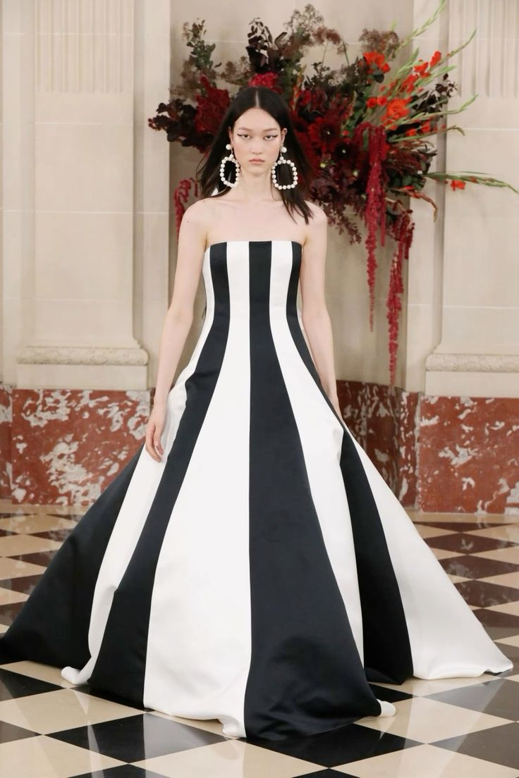 a woman in a black and white striped wedding dress stands on a black and white checkered floor. she's looking at the camera. there is a large bouquet of red flowers behind her.