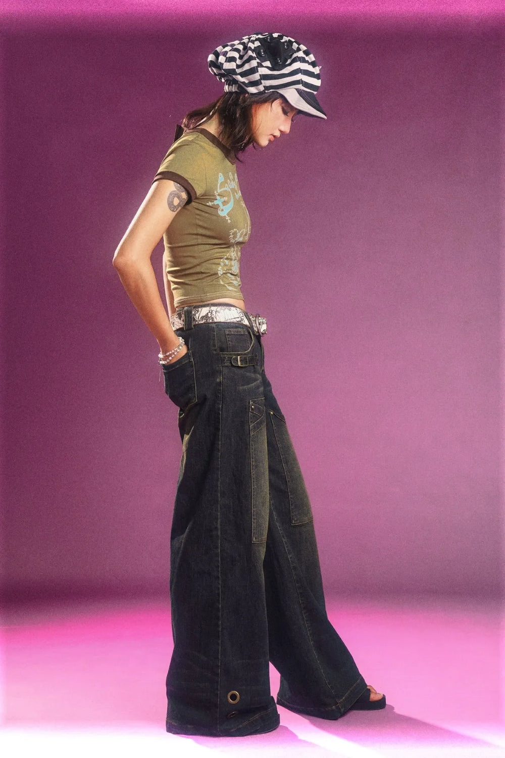 a fulllength side view of a young woman posing in a studio against a pink background.  she's wearing a  loosefitting olive green tshirt with a graphic design, lowrise dark wash wideleg denim pants with cargo pockets and a silver belt, and a black and white striped newsboy cap.  her pose is relaxed, hands in her pockets, and she's wearing what appears to be dark sandals. the overall style is reminiscent of 2000s fashion.