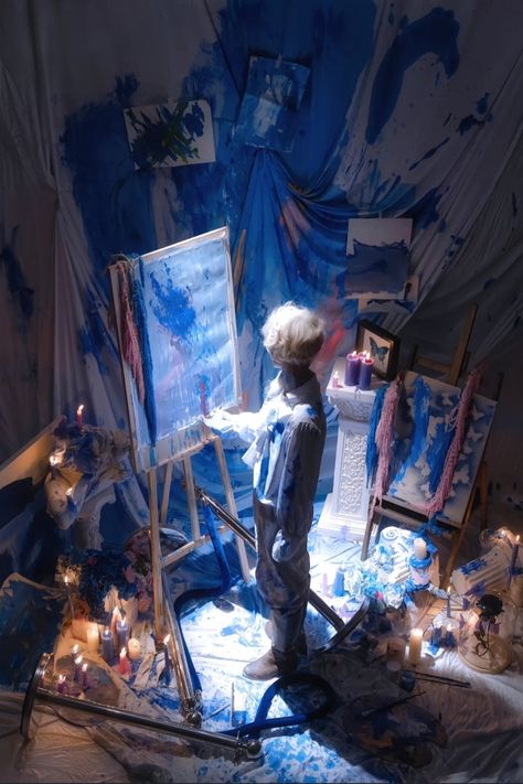 a man with blond hair, dressed entirely in white, stands amidst a chaotic art studio, entirely awash in blue paint. he intently paints on a canvas, illuminated by a spotlight that casts his figure into sharp relief against the vibrant blue backdrop. the scene is viewed from a high angle, emphasizing the immersive nature of his artistic process.  candles and paintsplattered objects contribute to the overall atmosphere of passionate, messy creation.
