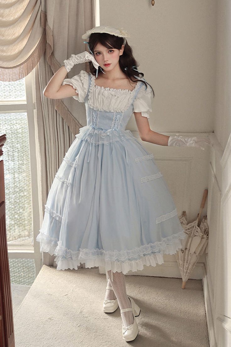 a young woman poses in a light blue and white lolita style dress. the dress features a square neckline, puff sleeves, and a flowing skirt with lace detailing. she is also wearing white gloves, a white hat, and white shoes. the setting appears to be indoors in a welllit room with a vintage feel.  the soft lighting creates a dreamy and romantic atmosphere.