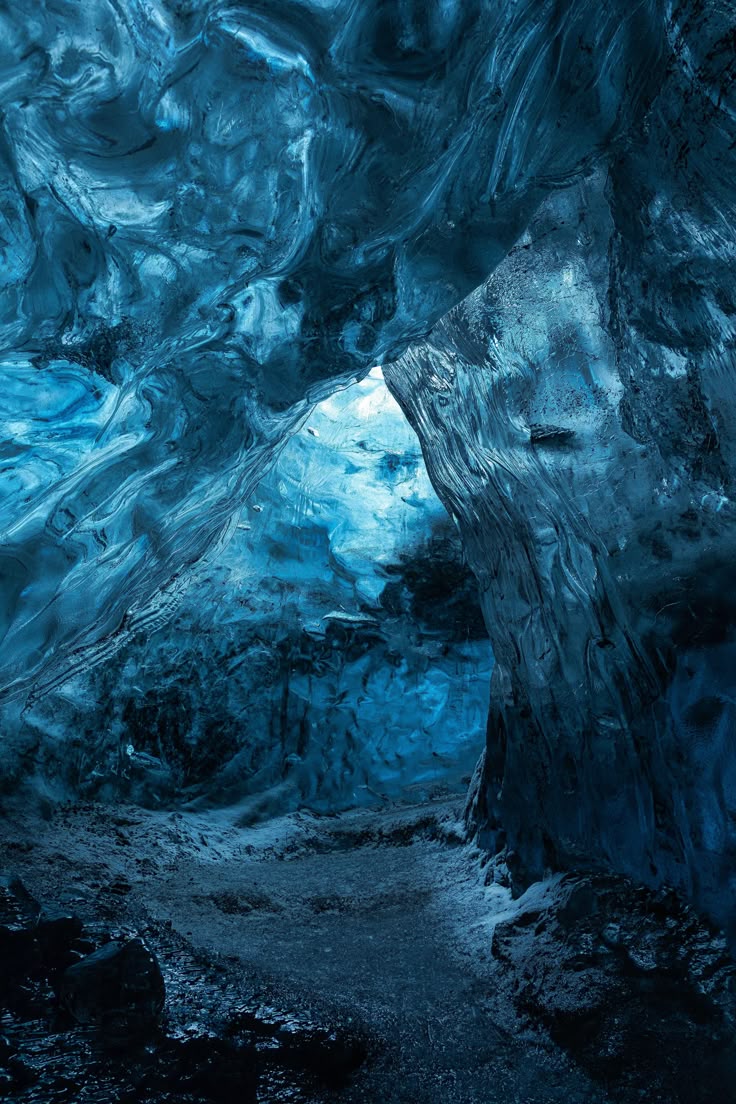 the image shows the interior of an ice cave. the ice is a deep blue color and appears to be part of a glacier. natural light streams into the cave from an opening, highlighting the intricate formations of the ice. the cave floor is rocky.