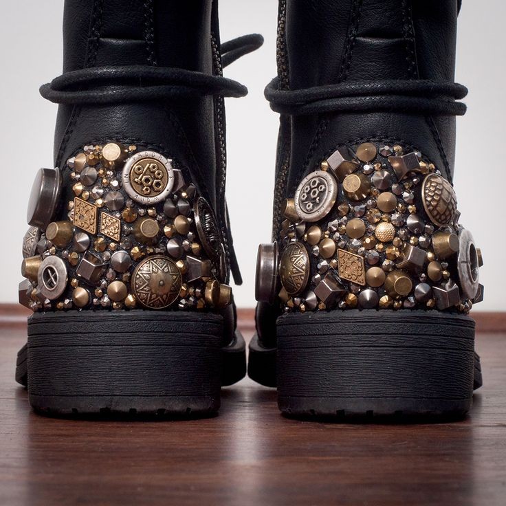 a closeup, back view image of a pair of black leather boots heavily embellished with various metal and crystal studs, buttons, and rivets in gold and silver tones.  the boots are set against a dark brown wooden surface, and the intricate detailing of the embellishments is clearly visible. the boots have a chunky, somewhat rugged appearance.