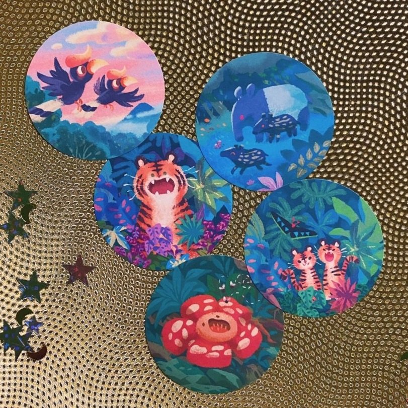 the image shows six round stickers or coasters featuring whimsical illustrations of jungle animals and plants on a gold textured background.  each sticker depicts a different scene: a pair of hornbills in flight, a family of tapirs, a roaring tiger, two tiger cubs, and a large, vibrant red flower. the artistic style is colorful and cartoonish. the stickers appear to be designed for decorative or stationery purposes. some small starshaped decorations are also visible around the stickers on the gold surface.