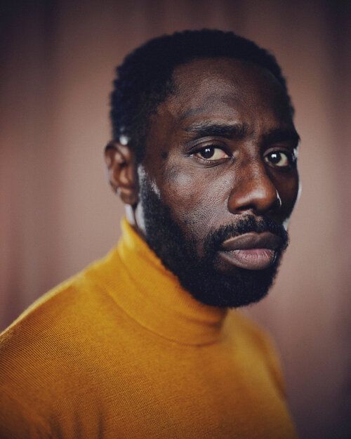 a portrait of a black man with short black hair and a beard, wearing a mustard yellow turtleneck sweater, against a blurred brownish background. he is looking directly at the viewer. the photo appears to be taken indoors with natural lighting.