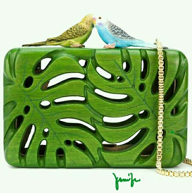 a purse or clutch bag with a monstera leaf design and two budgerigars on top. the purse is green with holes cut out in the shape of monstera leaves, exposing the tan interior. on top, there's a yellowgreen bird and a blue bird, touching beaks. a gold chain is attached on the right side of the purse. the white background highlights the purse and its elements. in the bottom right corner, there is a green signature.
