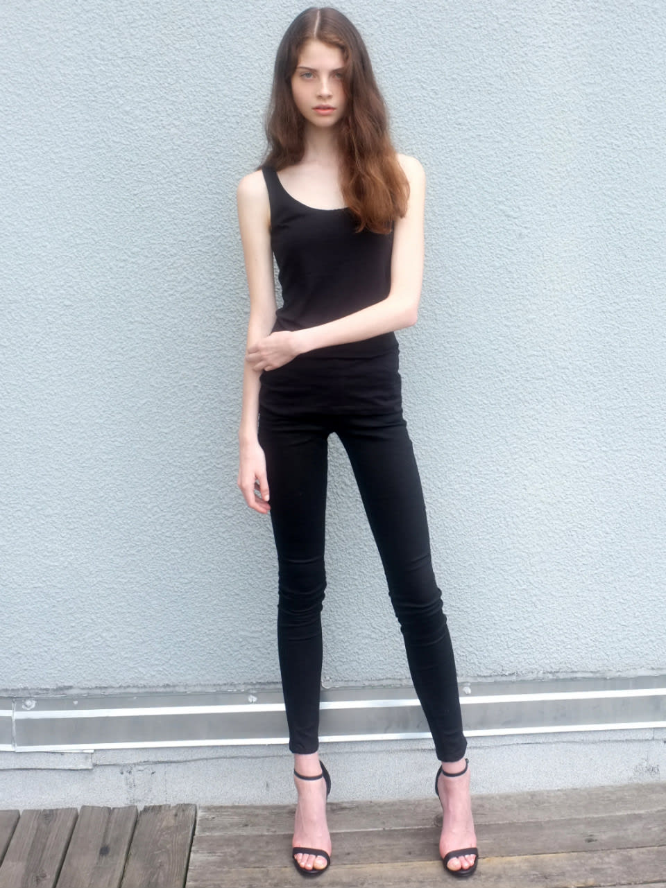 a fullbody shot of a young, slender woman standing in front of a light gray, textured wall. she is wearing a black tank top, black skinny jeans, and black high heels with ankle straps. her long, brown hair is down and she is posing with one hand resting on her hip. she is standing on a wooden platform.