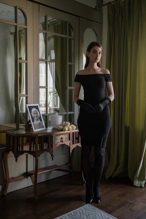 a young woman in a black offtheshoulder dress and black gloves stands solemnly in front of an antique console table.  a framed portrait of an older woman sits on the table, along with a small bouquet of white flowers. the setting is a dark, elegant room with vintage furniture, large windows, and heavy curtains. the overall mood is somber and suggestive of a funeral or mourning. the woman's posture and expression convey a sense of sadness and grief. the image is styled with a focus on creating a mood of elegance and melancholy.