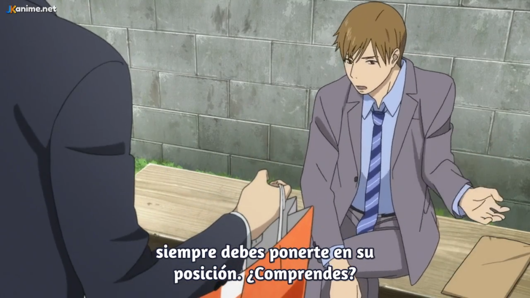 the image shows a scene from an anime with two men sitting on a bench, engaged in a serious conversation. the man on the left, wearing a dark suit, is handing a bag to the younger man on the right. the younger man, wearing a grey suit, is listening attentively with a concerned expression. the background features a plain wall, and the overall lighting suggests a daytime setting. the scene conveys a sense of mentorship or guidance being offered from the older man to the younger one.