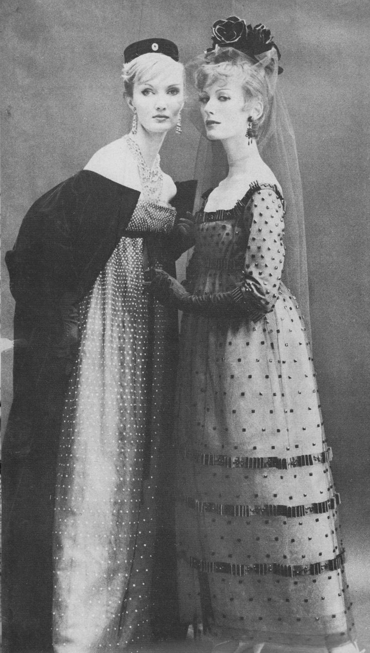 two women stand sidebyside in a black and white photograph, showcasing vintage fashion. the woman on the left wears a floorlength gown with a dense pattern of small dots and a dark shawl draped over her shoulders. she also has a small, flat hat and statement jewelry. the woman on the right wears a dress with a similar polka dot pattern, featuring bands of embellishment around the skirt. she is adorned with a rose headpiece and a sheer veil. both women have elegant poses and look directly at the camera.