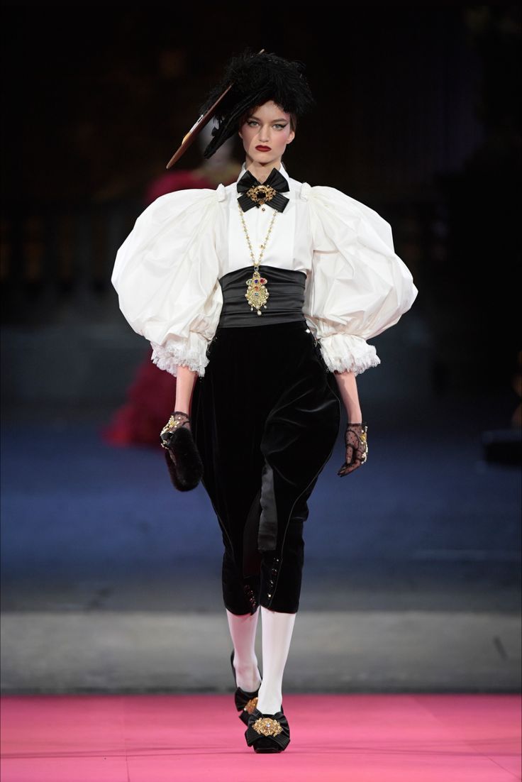 a model walks down a pink runway wearing a stunning haute couture outfit.  the ensemble features a dramatic white blouse with large, puffy sleeves, a black velvet highwaisted pant, and a black velvet bodice.  she accessorizes with a statement gold and black necklace, a black bow tie, black gloves, and ornate black shoes. her hair is styled up with a dark hat. the overall look is elegant, sophisticated, and dramatic. the background is dark and blurred, focusing attention on the model and her attire. the photo appears to be from a fashion show.