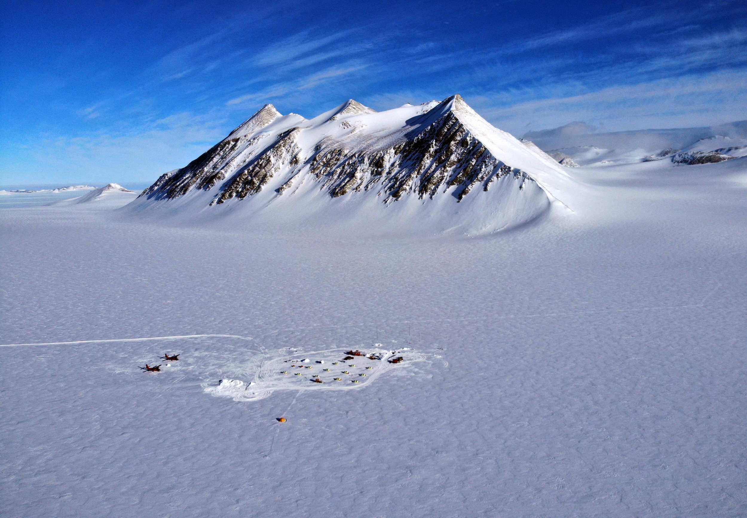 a breathtaking aerial view of a snowcovered mountain range in antarctica. the mountains, rising dramatically from a vast, white expanse of ice and snow, create a stark and beautiful landscape. the clear blue sky and the sun reflecting off the snow create a sense of serenity and isolation. in the foreground, a small research station or camp is visible, highlighting the human presence in this remote and unforgiving environment. the image captures the majesty and grandeur of the antarctic wilderness, a place of extreme cold, harsh conditions, and incredible beauty.