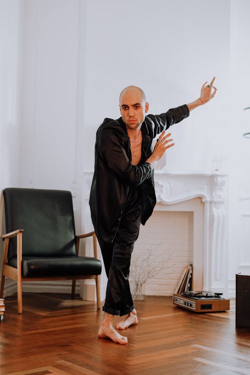 a bald man wearing a black satin pajama set is captured middance in a minimalist interior. he is gracefully poised, showcasing expressive movement. the setting includes a wooden floor, an armchair, a fireplace, and a record player, adding to the overall atmosphere of artistic contemplation and personal style. the image conveys a sense of elegance and serenity, focusing on the individual's expressive dance and artistic sensibility within a comfortable domestic space.