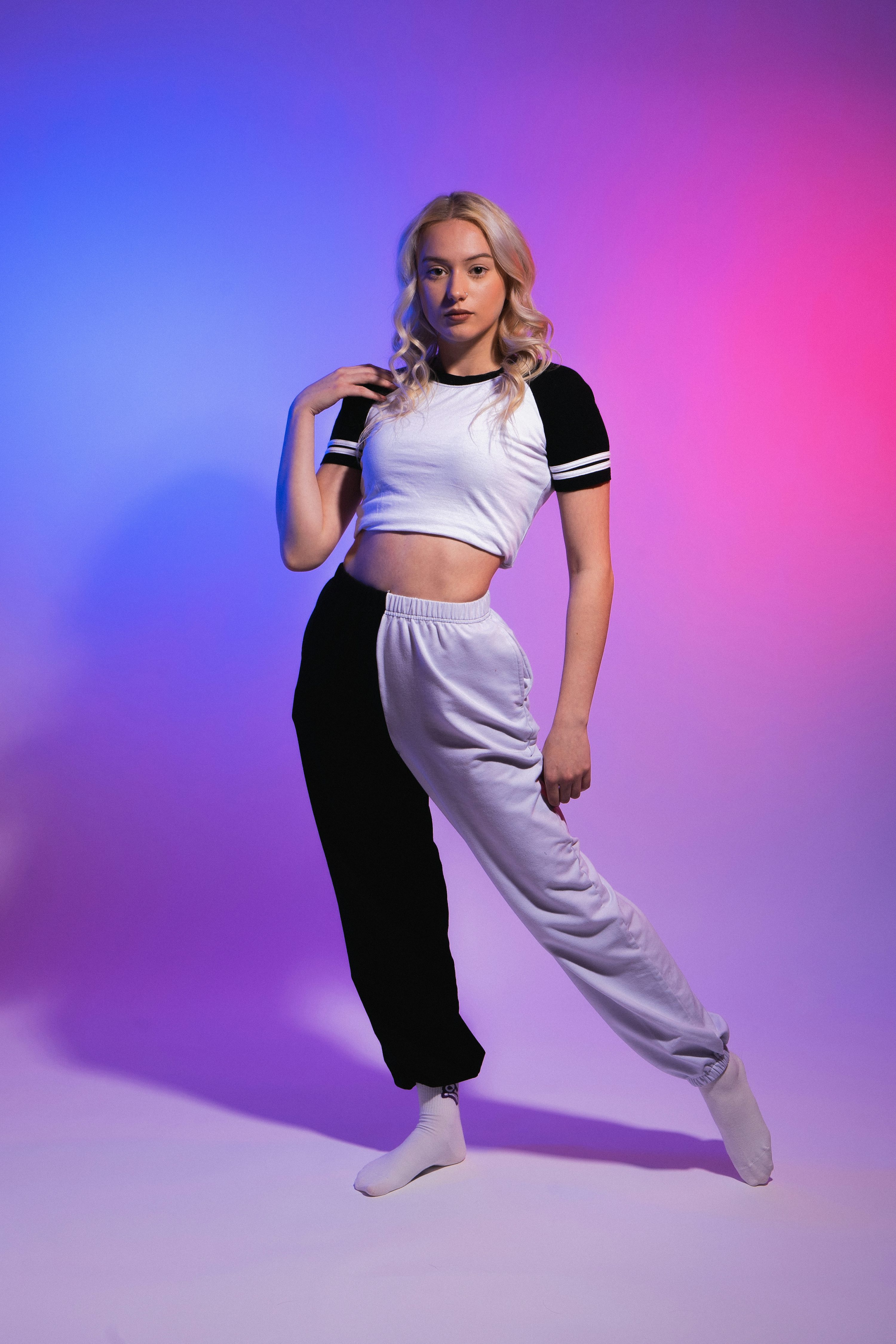 a young woman with blonde hair is standing in a studio against a vibrant purple and blue background. she's wearing a white and black twotone tracksuit consisting of a cropped white top with black sleeves and split sweatpants, one side black and the other white. she's striking a confident pose, slightly angled, with one leg extended. her style is sporty and casual, and the image's neon lighting enhances the trendy vibe.