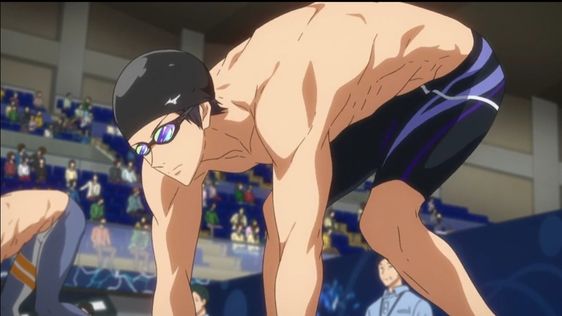 the image shows a male swimmer in a starting position on a starting block. he is wearing blue swim trunks and goggles and is positioned in a crouched stance,  with his body turned towards the viewer and his head looking down. the background shows a blurry indoor swimming pool setting with spectators in the stands.