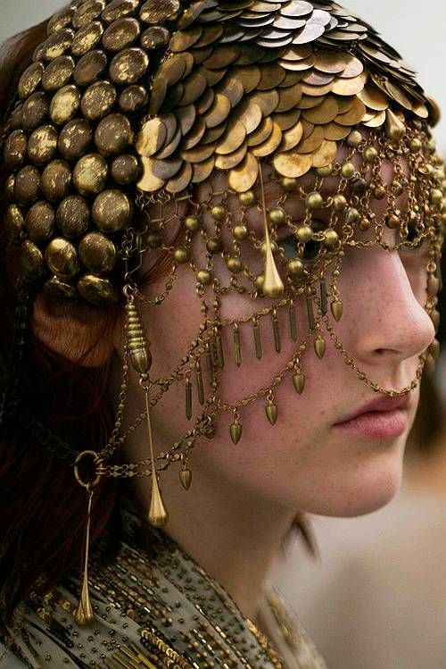 a woman's face is seen in profile, adorned with an elaborate gold headpiece. the headpiece features a dense layer of gold coins covering the top of the head, with intricate chains and dangles cascading down around the face. the jewelry has a vintage or antique appearance, with a rich, warm tone. the woman has fair skin with noticeable freckles. her expression is neutral.