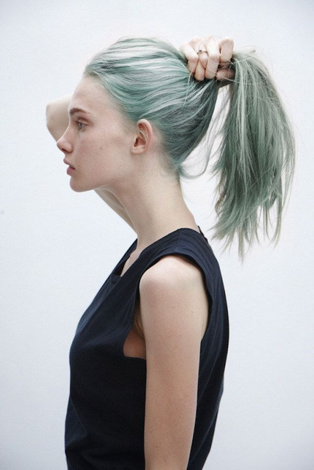 a woman with long, dark green hair pulled back into a ponytail is wearing a black tank top, looking away from the camera, against a white background.