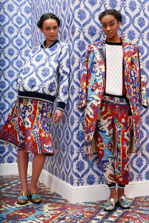 two models are posing against a wall with blue and white patterned wallpaper. both are wearing brightly colored clothing with intricate patterns reminiscent of rugs or tapestries. the model on the left wears a white top with blue and white patterns and a red, blue and yellow patterned skirt. the model on the right wears a matching pantsuit with a cream colored sweater.  the floor is also covered in a similar patterned carpet.  both women are wearing gold and blue tassel loafers.  the overall style is vibrant and bohemian.