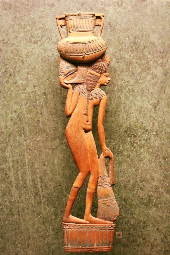 the image shows a wooden statue of an egyptian woman carrying a vase on her head and a bag in her hand. the statue is carved in profile and shows the woman's nude body. she stands on a rectangular base decorated with carved lines. the statue is displayed against a green marble background.