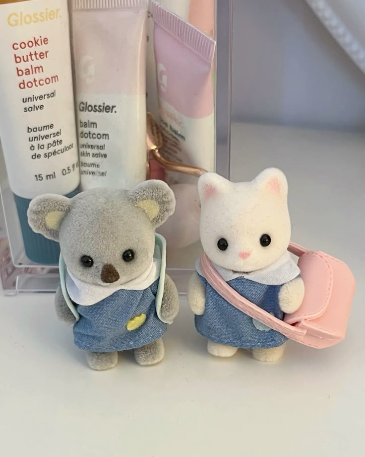 the image features two sylvanian familiesstyle toys, a koala and a cat, positioned in front of several glossier balm dotcom tubes. the koala, dressed in a denim dress with a small yellow patch, sports a miniature backpack, while the cat, also in a denim dress with a white collar, carries a pink miniature purse. behind them are tubes of glossier balm dotcom in various flavors, including cookie butter, arranged within a clear container. the scene is brightly lit, emphasizing the pastel aesthetic of the products and toys.