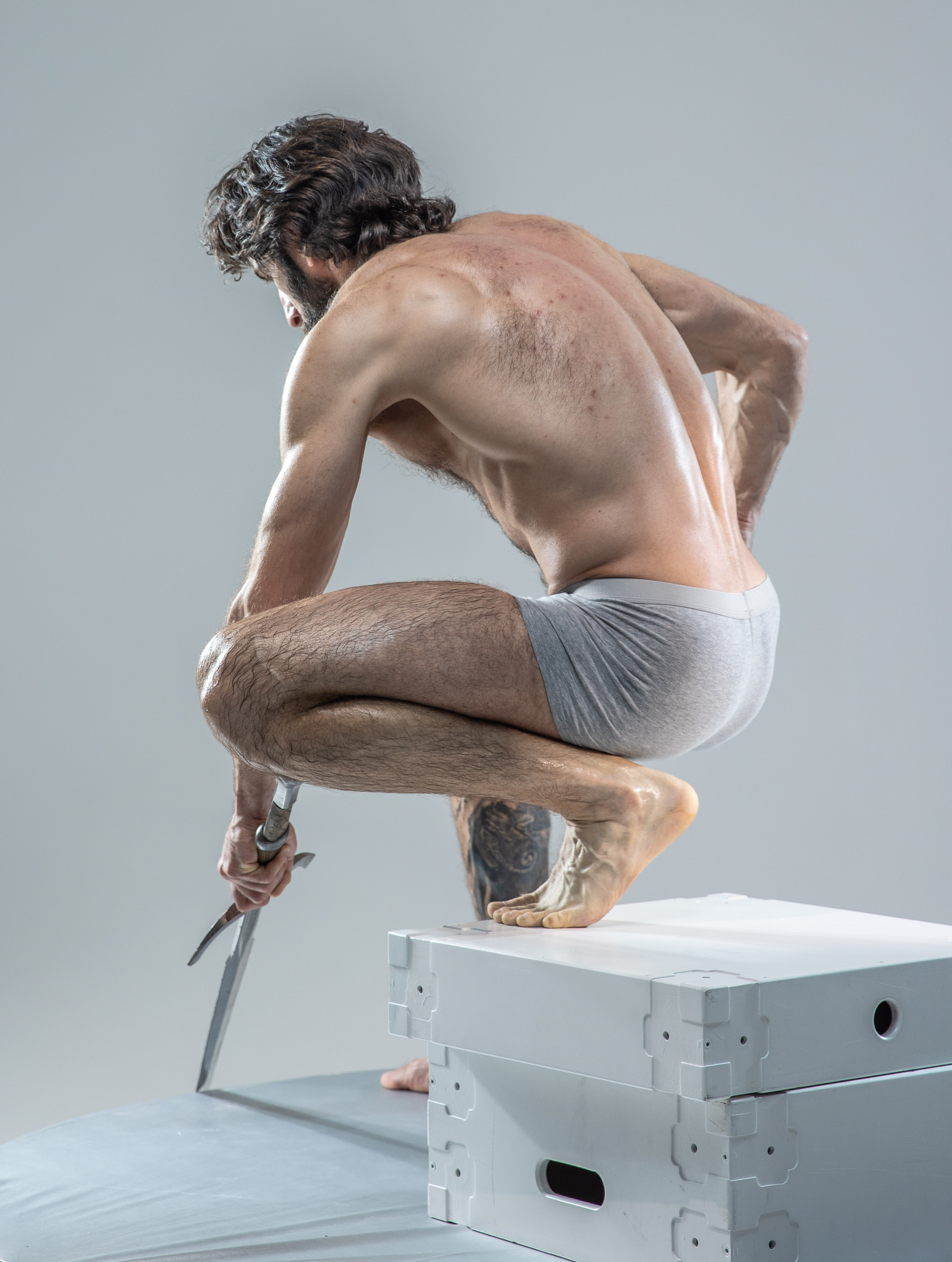 a muscular man with dark hair and beard crouches on a white cube in a dynamic pose. his back is to the camera, highlighting his welldefined musculature. he holds a sword in his left hand, pointing it downwards, suggesting impending action. the scene is set against a simple grey studio backdrop, lit with even, bright lighting that accentuates his physique.