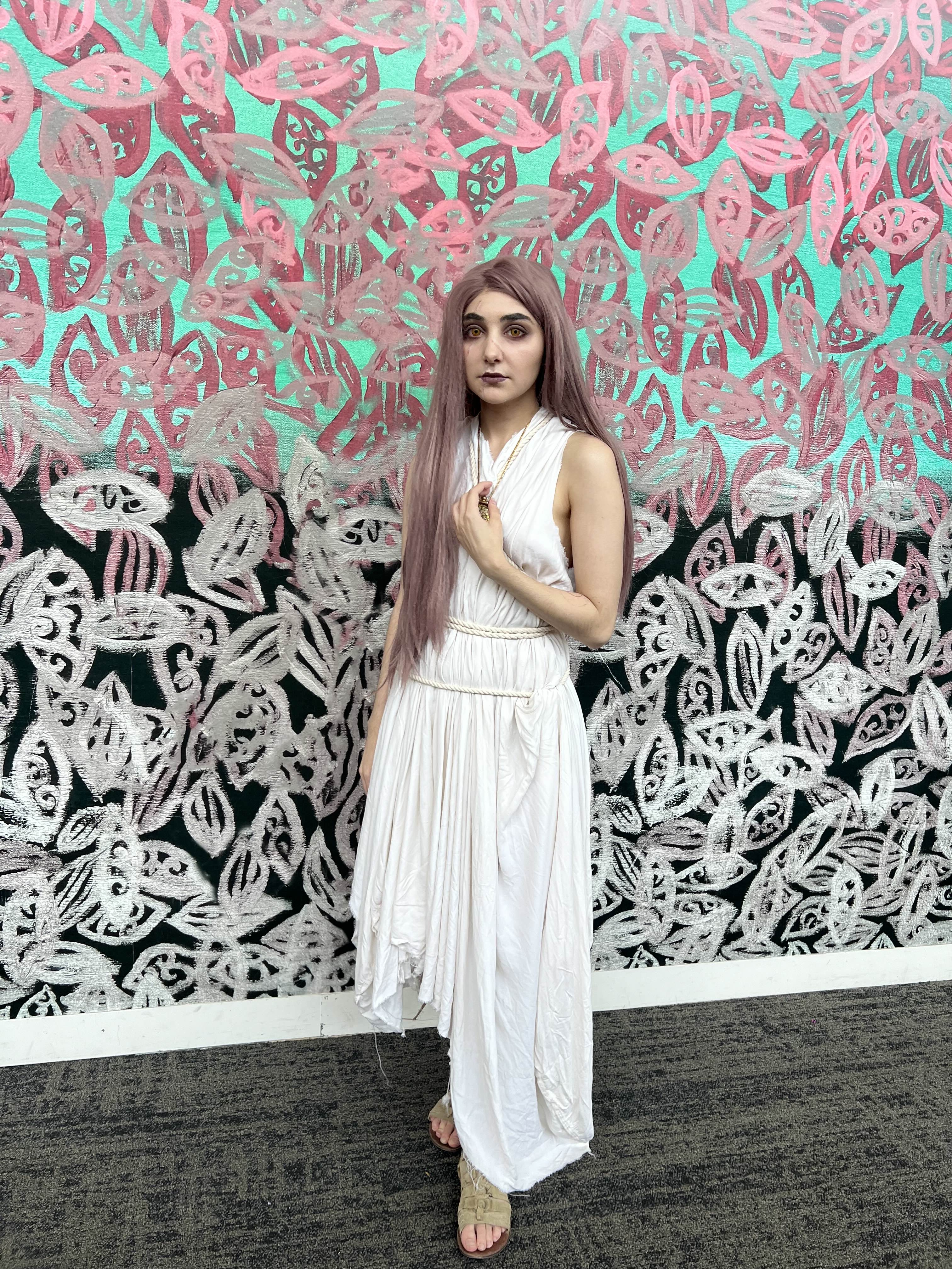a fullbody shot of a young woman with long pink hair standing in front of a vibrant wall covered in abstract art. she's wearing a flowing, asymmetrical white maxi dress with a simple design, accessorized with a delicate necklace and sandals. the artwork behind her features a repeating pattern of stylized leaves in shades of pink and white on a teal background, creating a striking contrast with her dress. the overall mood is artistic and fashionable.