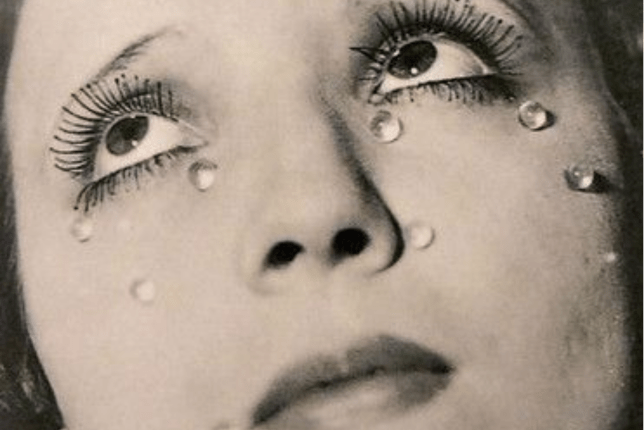 a closeup black and white photograph of a woman's face, possibly a portrait. her eyes are accentuated with dark eyelashes and makeup, and small, round objects resembling teardrops are strategically placed on her cheeks. the overall mood is melancholic and artistic, suggestive of a surrealist style.  the image's style and composition is reminiscent of man ray's work.