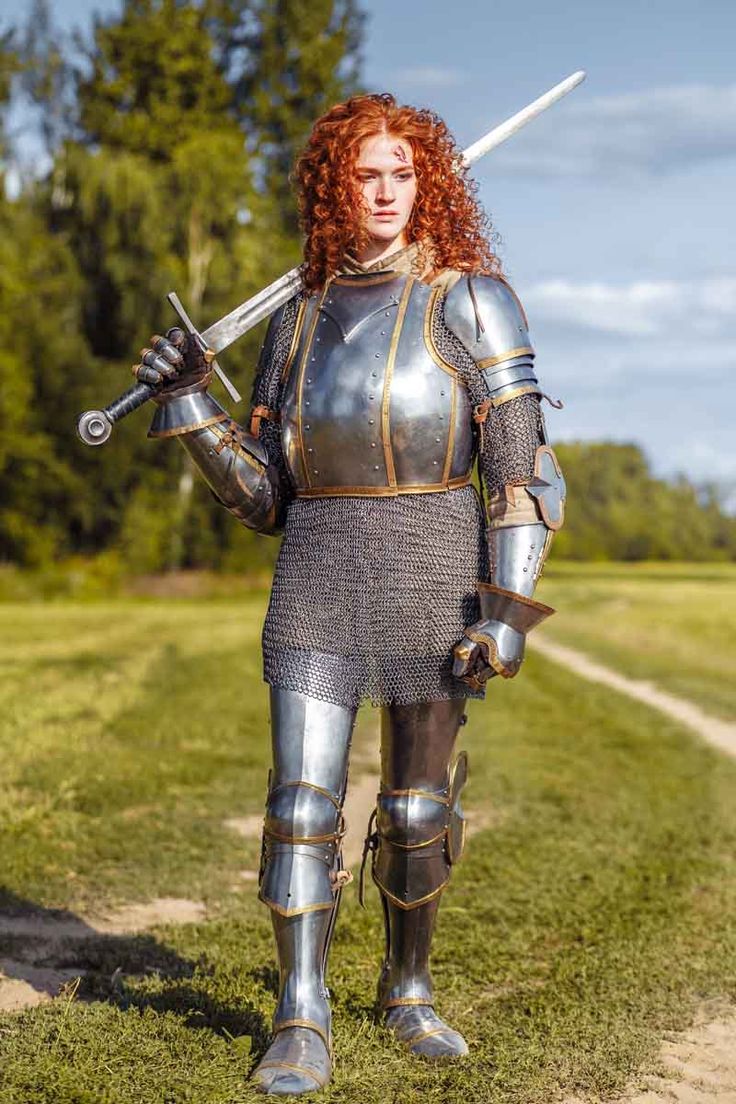 the image showcases a female knight in full medieval plate armor and chainmail. she has vibrant red, curly hair and is carrying a longsword across her shoulder. the knight stands in a grassy field with a dirt path leading away from her. her armor is detailed, showcasing metallic highlights and intricate designs. she appears strong and confident, portraying a powerful female warrior in a historical or fantasy setting. the image is welllit, emphasizing the details of her armor and the scenery.