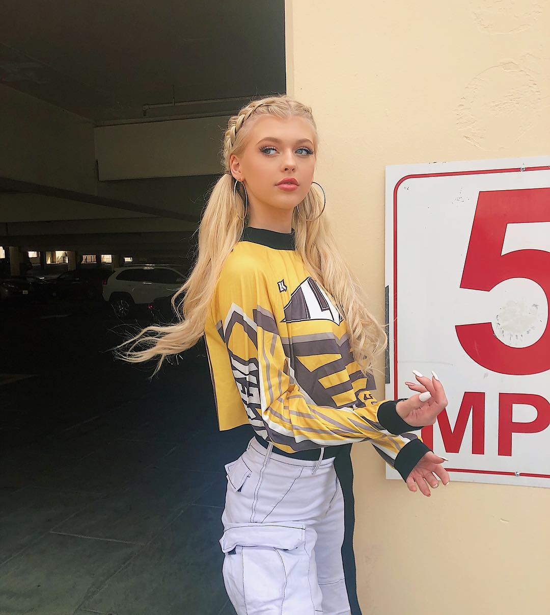 a woman with blonde hair styled in pigtails and braids stands in front of a parking garage, wearing a yellow shirt and white pants. she is wearing hoop earrings and posing next to a speed limit sign that reads "5 mph". the background shows a parking garage with cars.