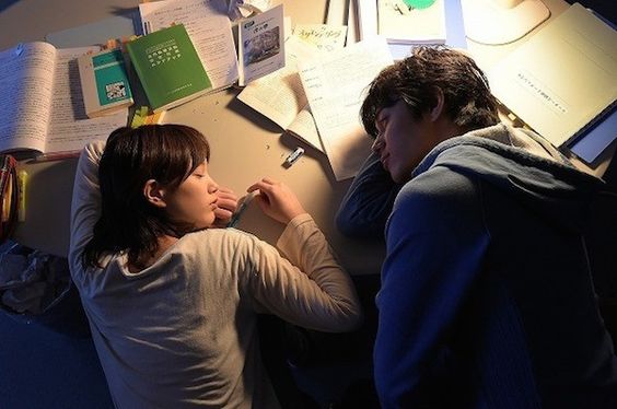 a young man and woman are sleeping on a table covered with books and papers.  they are indoors in a room lit by artificial light.