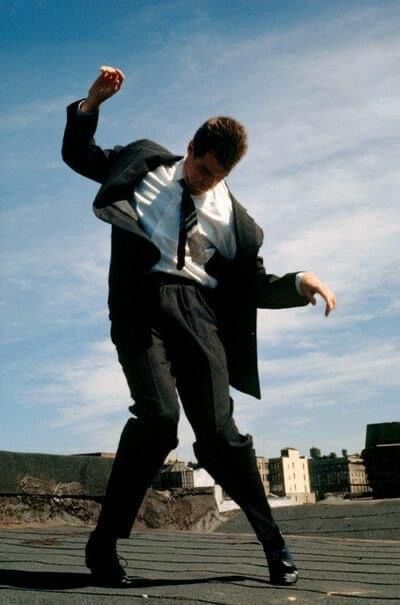 a man in a suit is energetically dancing on a rooftop, his arms flung out and his head tilted down. he is wearing black leather shoes, black pants, white shirt, and a black jacket with a loosened tie. in the background, a clear blue sky is dotted with white clouds and the tops of city buildings are visible. the low angle of the shot emphasizes the feeling of movement and freedom.