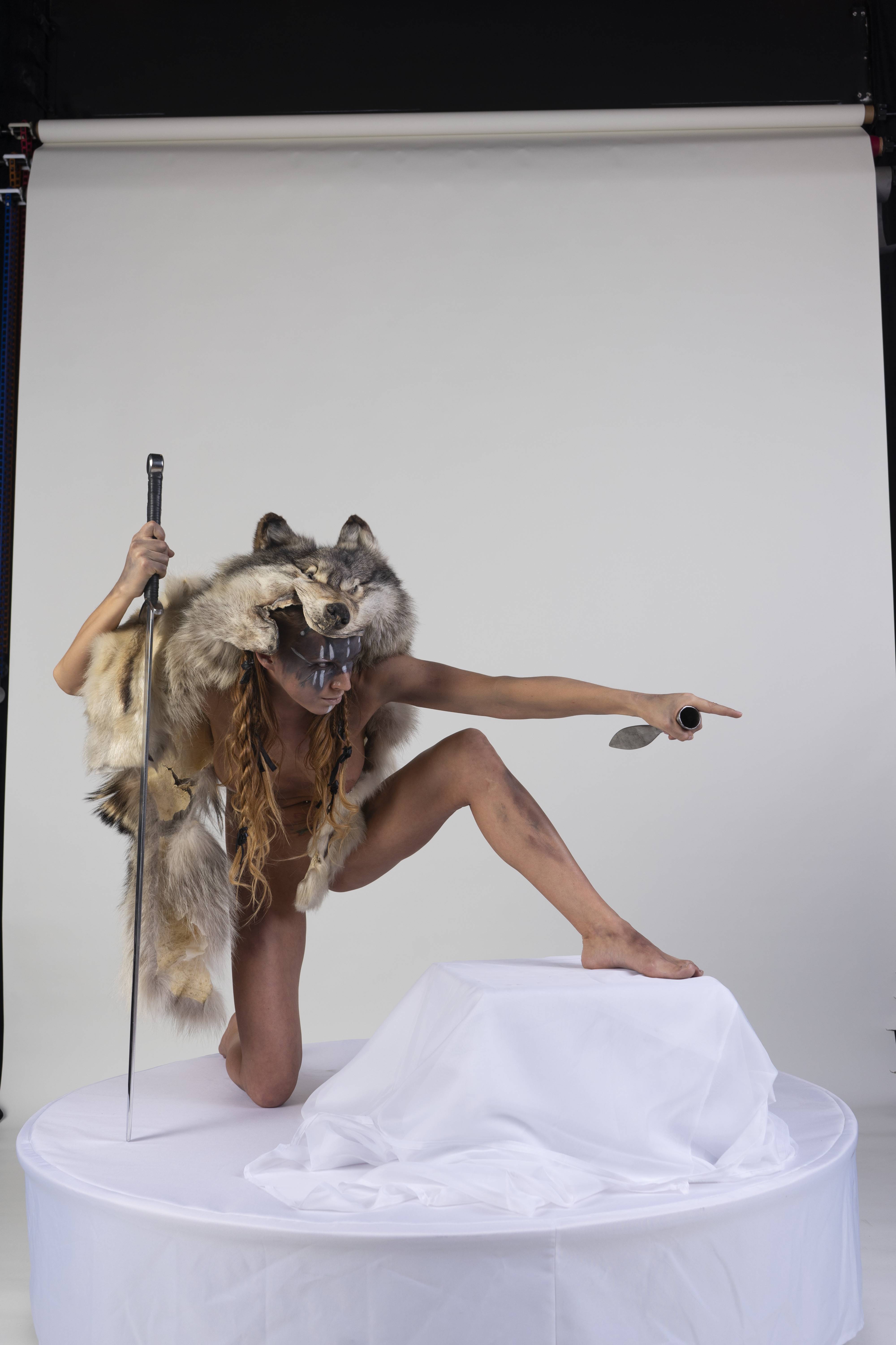a striking studio shot features a woman dressed in a tribal warriorlike costume. she is kneeling on a white surface, possibly a stage, with one knee on the ground and the other foot placed firmly on an elevated white draped platform. her shoulders are bare, and she is adorned with a headdress made from animal fur, specifically a wolf's head covering her forehead and extending down her back. her face is painted with tribal markings. in her left hand, she holds a sword, while her right hand is extended, pointing with a knife. the background is a neutral studio backdrop, highlighting the woman's striking pose and costume. the image conveys a sense of strength, tribal identity, and warrior spirit.