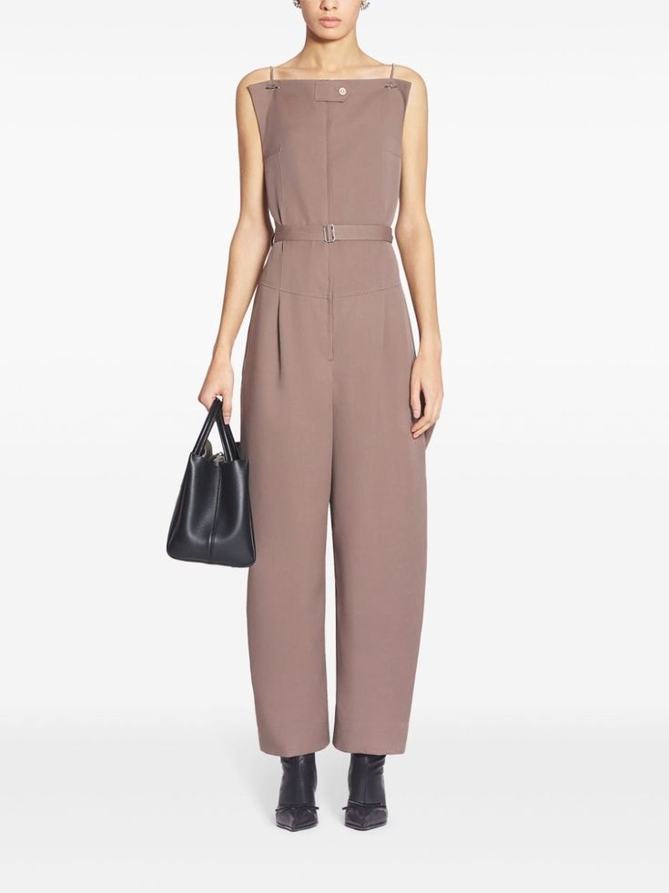 a woman wearing a simple brown jumpsuit, with a belt and a black handbag. the woman is standing in a studio setting against a white background. she is wearing black heels and looking at the camera.