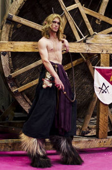 a man dressed in a fantasy costume stands posing in front of a large wooden wheel.  his costume includes a darkcolored tunic and pants with faux fur leg coverings that resemble goat legs and hooves. he has long blonde hair and is shirtless. he holds a whip and appears to be at a festival or fair. the background features rustic wooden structures.
