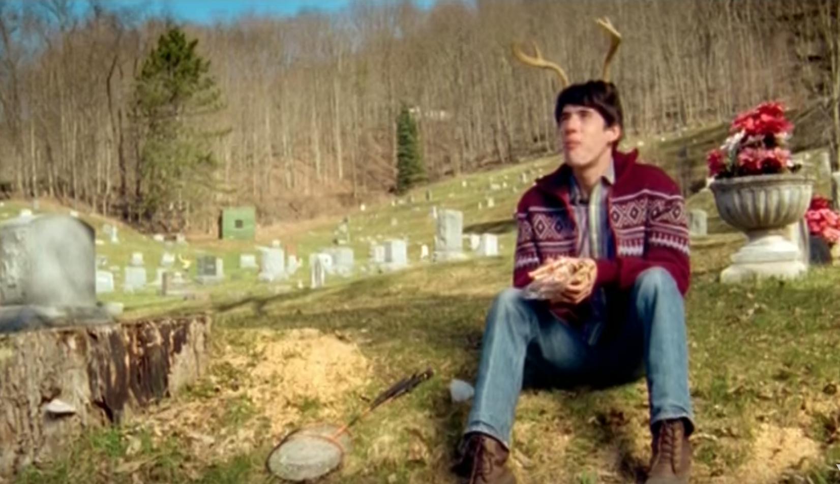 a young man with deer antlers on his head is sitting in a cemetery. he's wearing a red patterned sweater and jeans and is eating something from a bag. in the foreground, there is a badminton racket and what appears to be birdseed. the cemetery is located on a hill with a forest in the background. the image is brightly lit with natural sunlight, suggesting a sunny day.