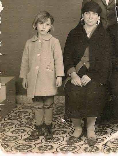 a vintage photograph shows a young girl standing next to a seated woman. the girl is wearing a lightcolored coat, pants, and boots with fur trim. the woman is wearing a dark shawl or coat, a hat, and shoes with embellishments. they are standing or sitting on a patterned floor. the photo has an old and worn appearance.