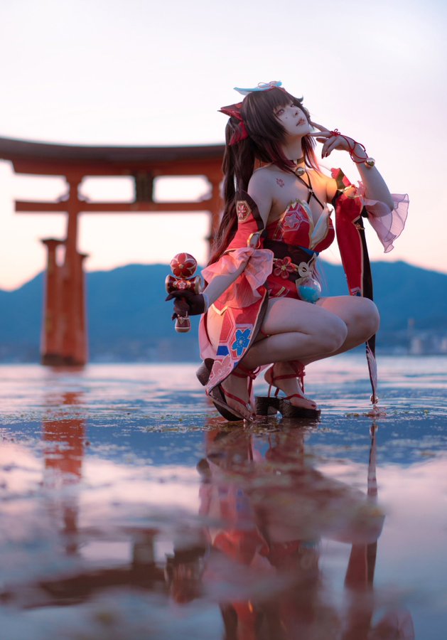 a woman dressed in an elaborate cosplay of the genshin impact character yae miko is posing in shallow water in front of a traditional japanese torii gate. the sun is setting in the background, casting a warm glow on the scene. the woman's costume is detailed and vibrant, featuring reds, pinks, and whites. her pose is both elegant and powerful. the reflection of the woman and the torii gate in the water adds a sense of depth and serenity to the image.